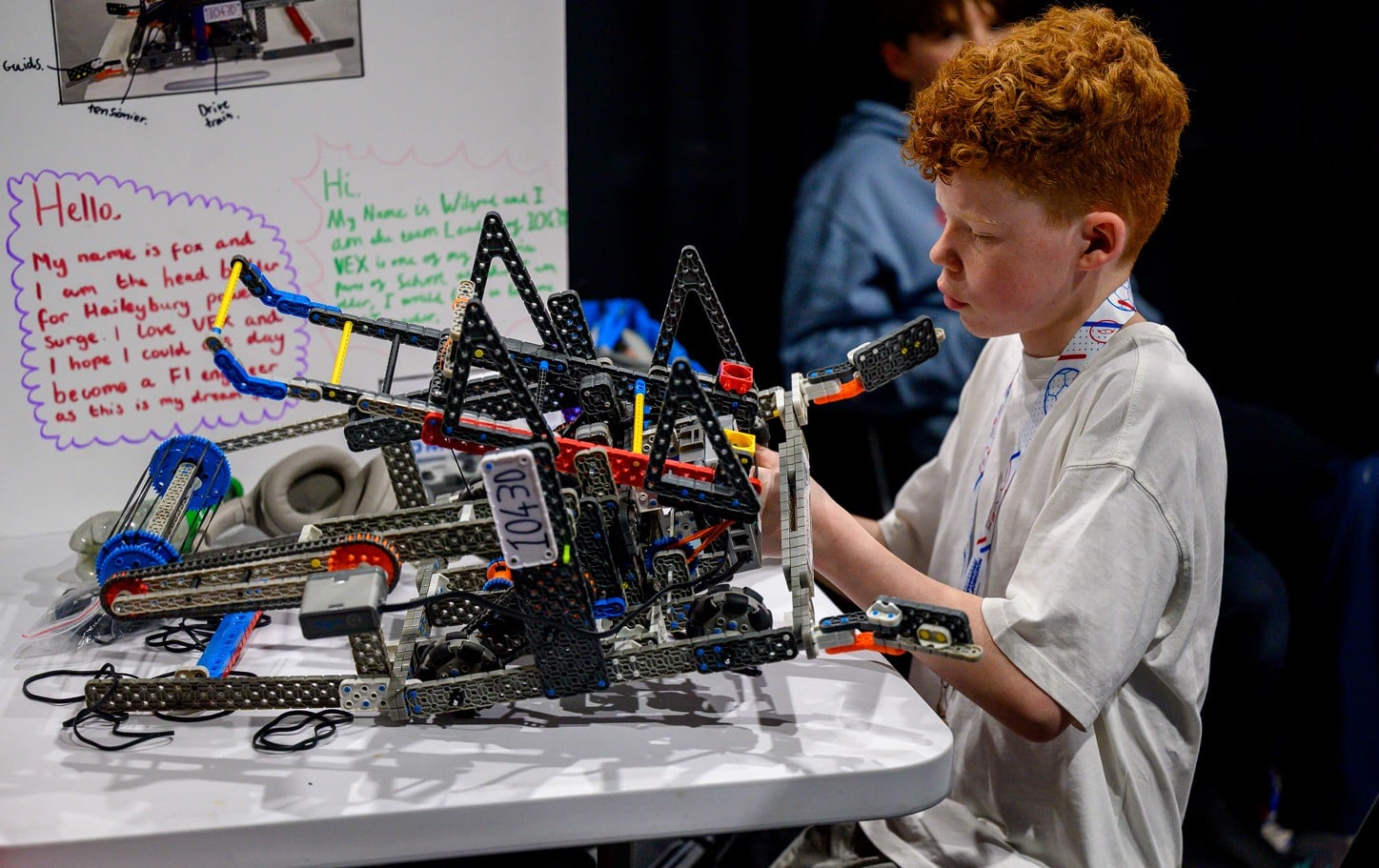 A student works on his VEX IQ robot at a pit table, with a handwritten team poster taped up behind him.