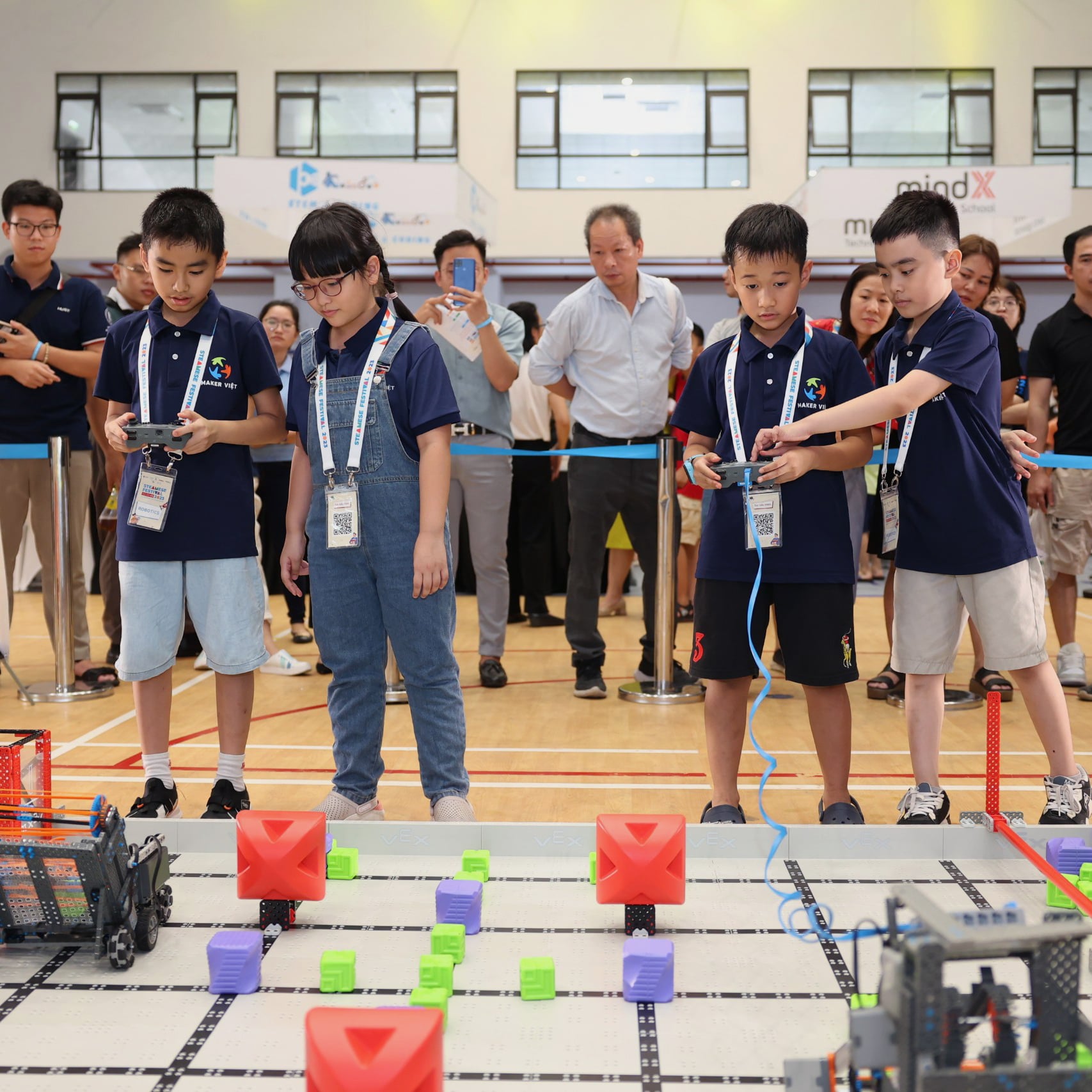 Four VEX IQ team members from two different teams drive their robots on the same field during a Teamwork Match, while spectators watch from behind a rope barrier.