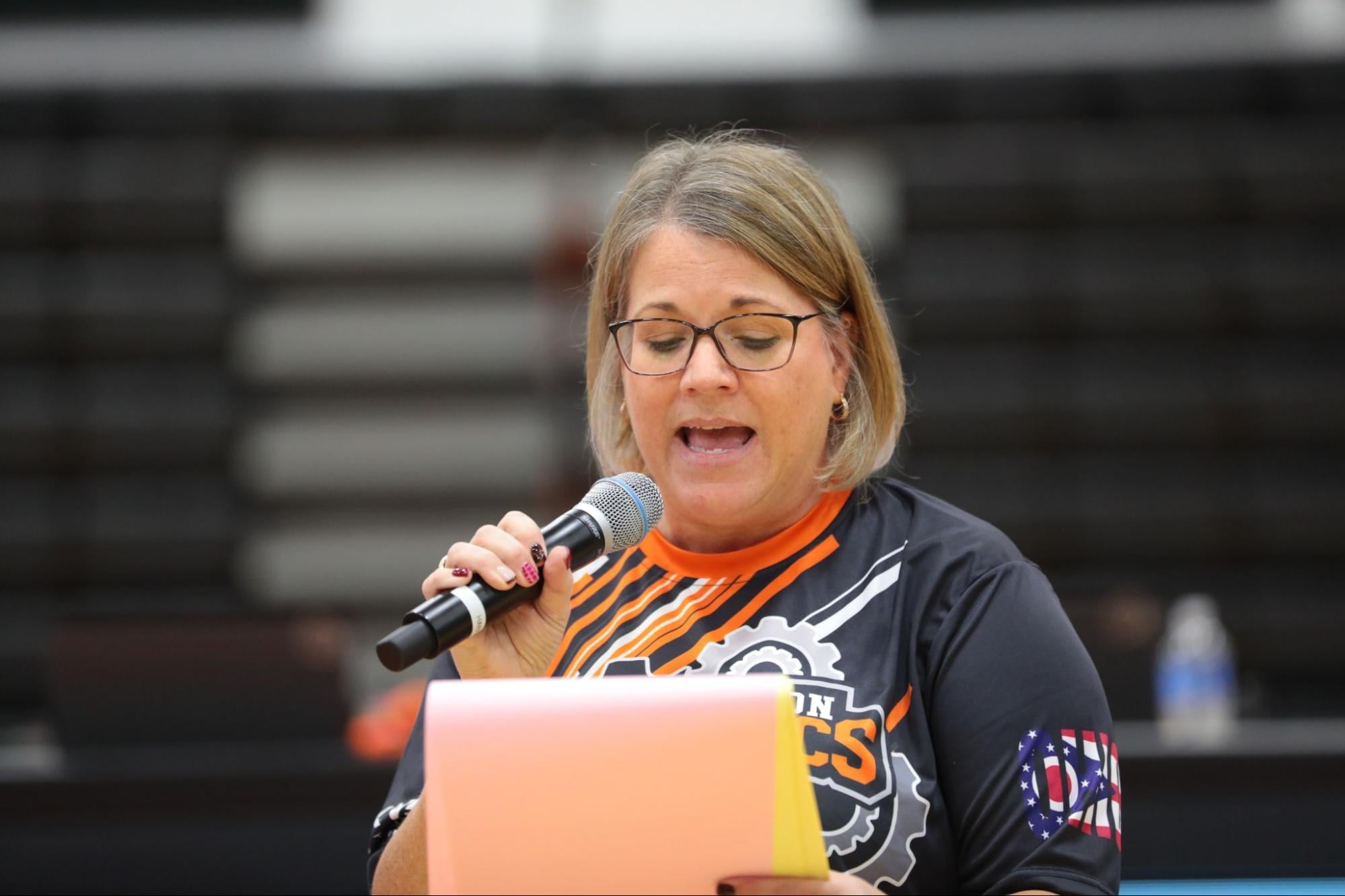 An Event Partner in a jersey speaks into a handheld microphone while reading from a folder in a gym.