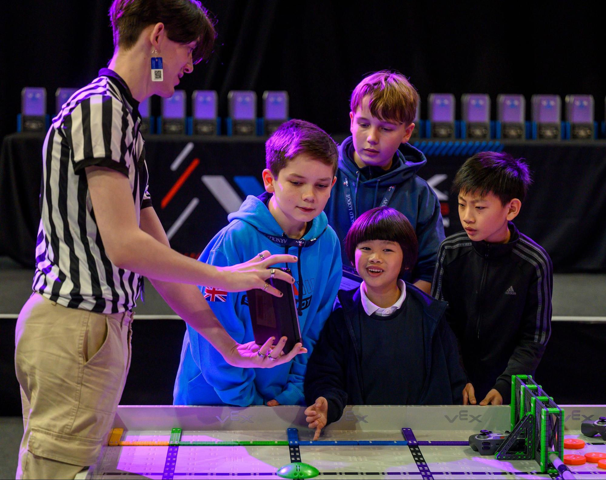 A referee holds out a tablet to show four VEX IQ team members the score at the end of their match.