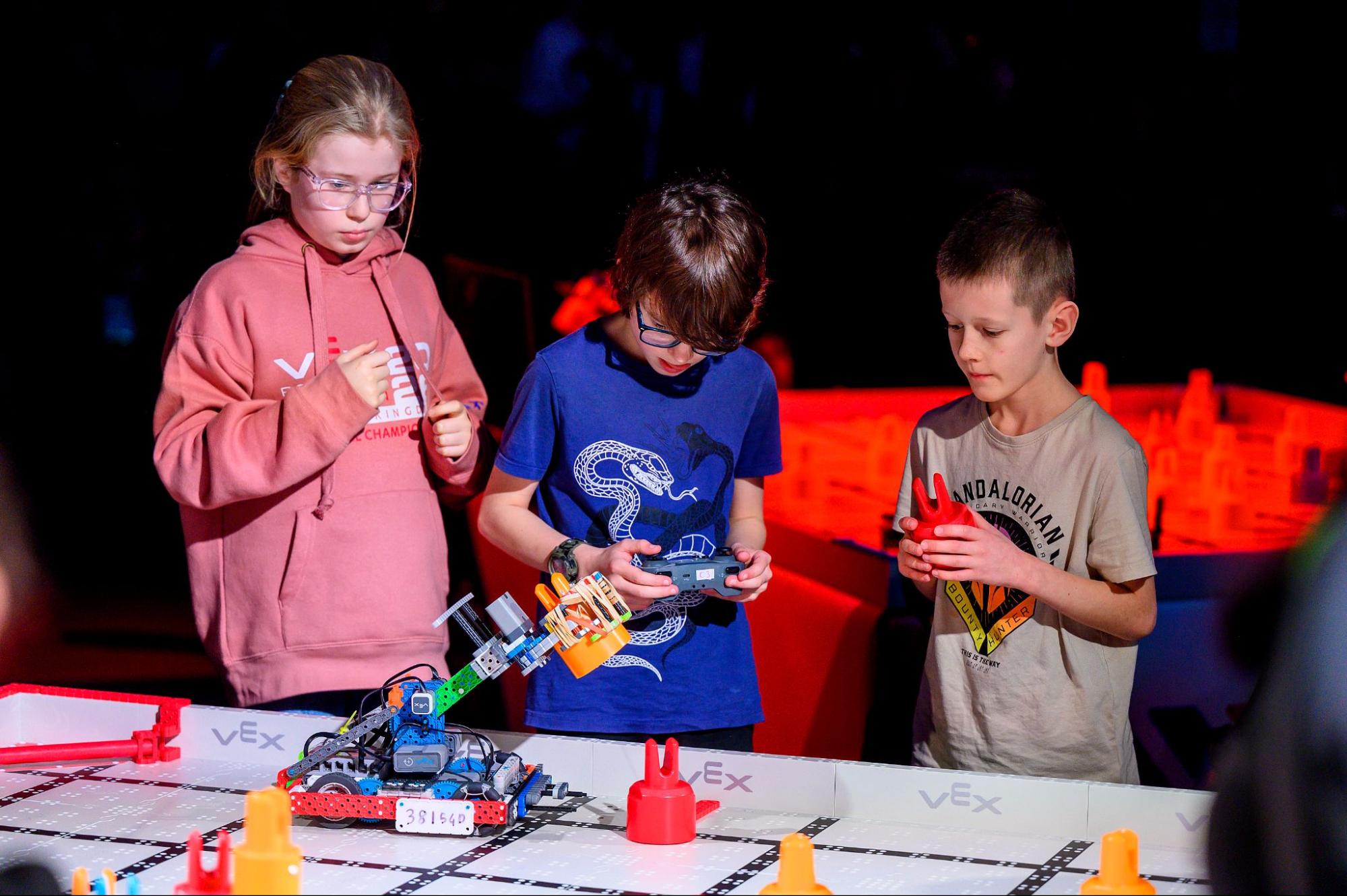 Three VEX IQ team members stand at their field as a match begins, one driving with a controller while a teammate holds a game piece ready to load.