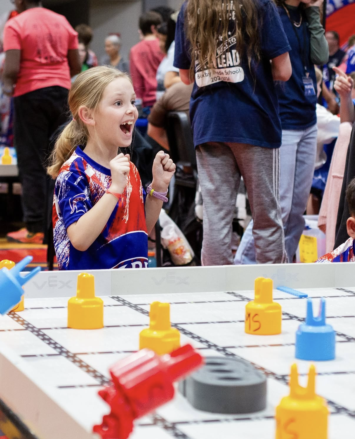 A young student in a team jersey cheers with both fists raised beside a VEX IQ field.
